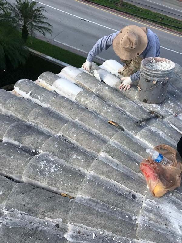 A worker wearing a tan hat and gloves repairs gray barrel roof tiles on a sloped roof near a road.