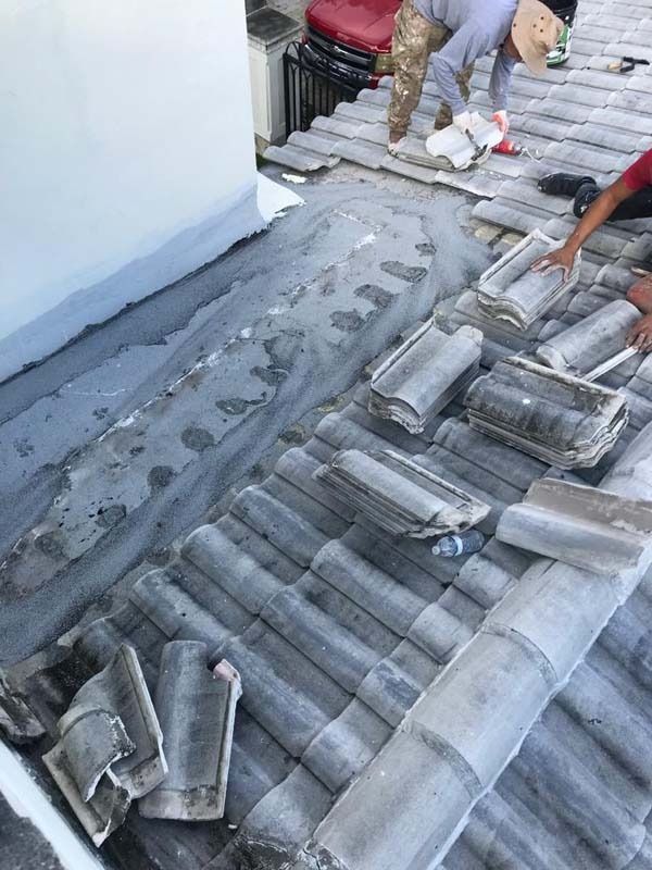 Workers repairing a grey tiled roof with a mortar patch against a white wall.