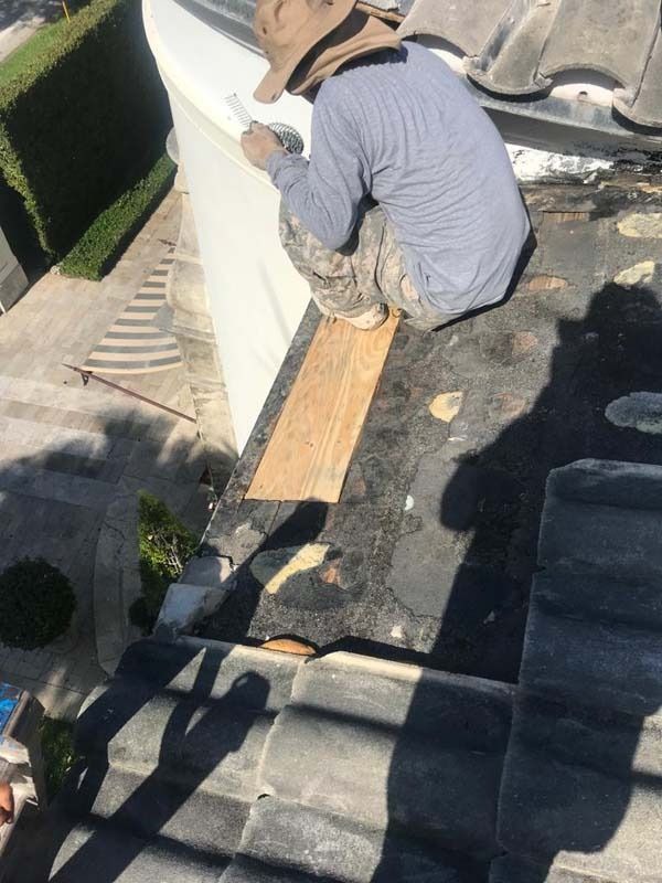 A worker in a grey long-sleeved shirt and hat squats on a tiled roof while applying sealant to the edge of a wall.