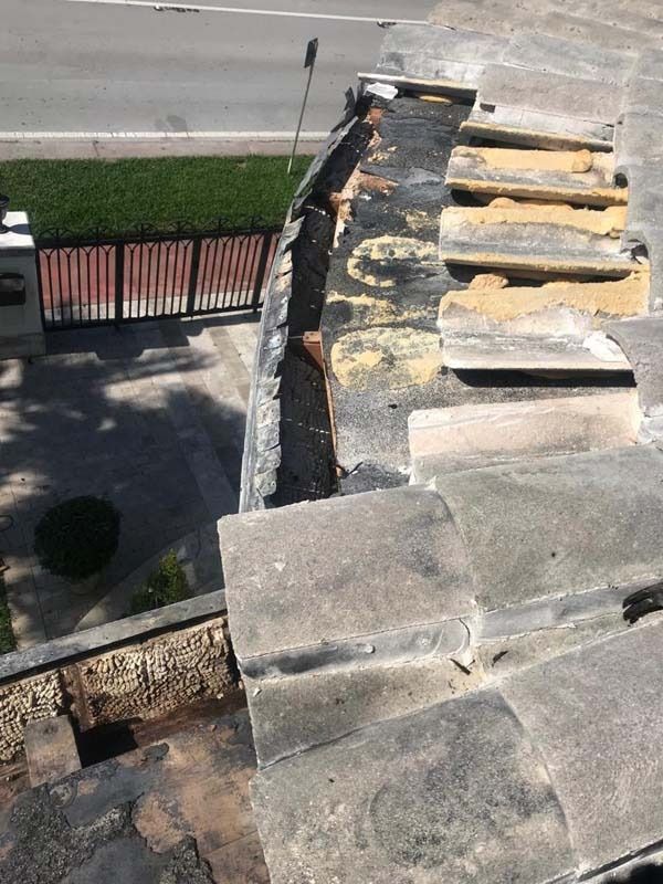A high-angle view of a roof under repair, showing missing stone tiles and exposed wood framing above a street.