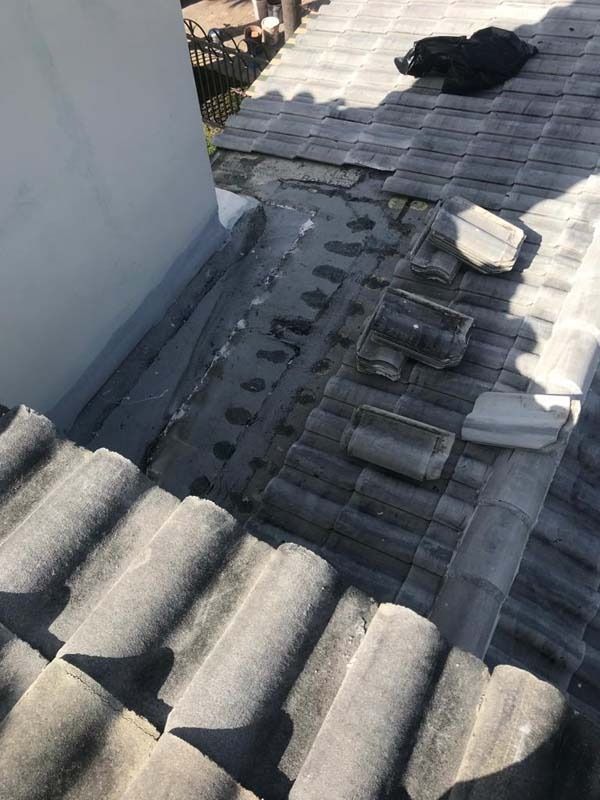 High-angle view of a tiled roof with several tiles missing, exposing the underlying underlayment near a light-colored wall.
