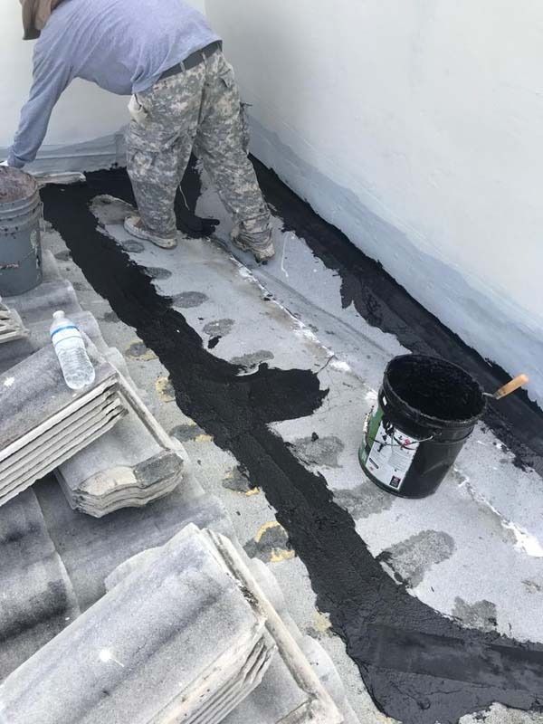 A worker applies black sealant along a joint on a gray tiled roof next to a bucket of roofing material.