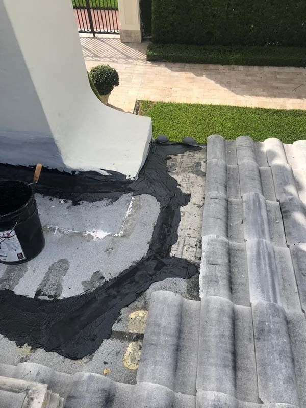Black roof sealant is being applied to a flat section of a tiled roof where it meets a white exterior wall.