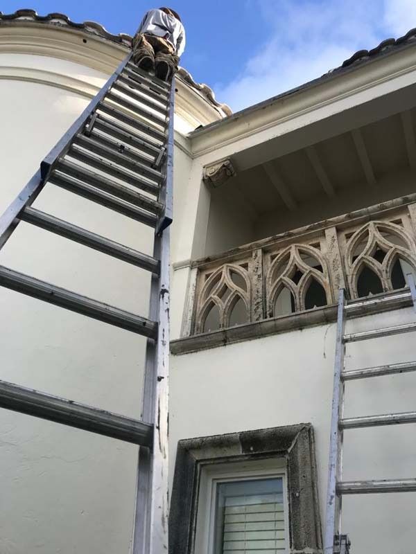 A person standing on an extension ladder propped against the roofline of a white building with an arched stone balcony.