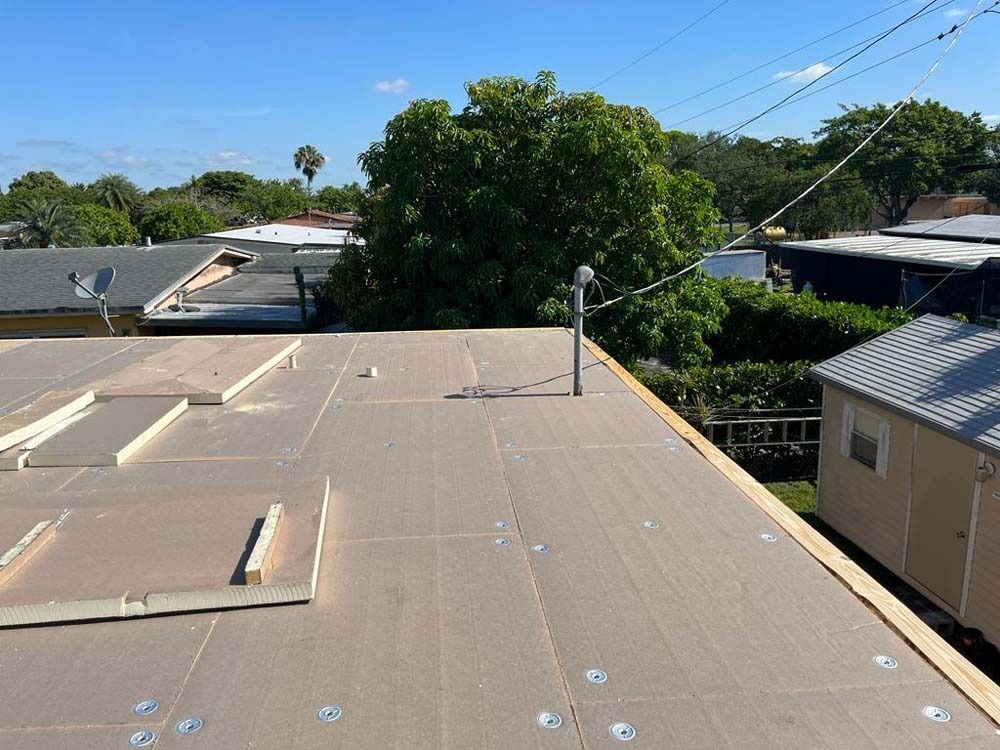 A flat roof covered in tan insulation boards, with metal fasteners, a vent pipe, and surrounding neighborhood trees.