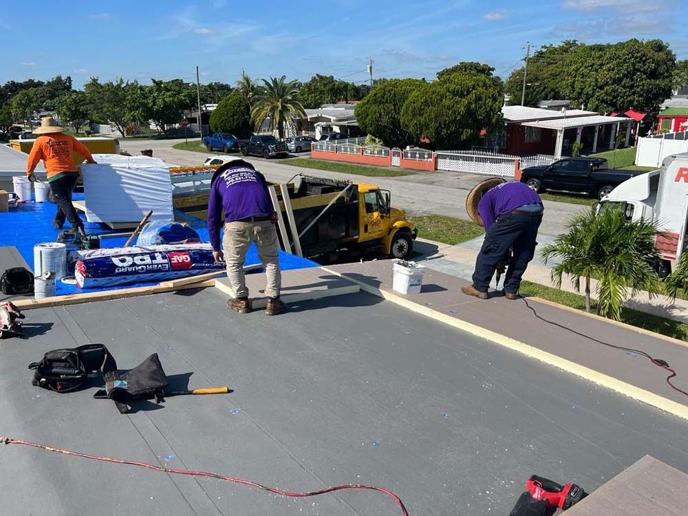 Construction workers installing a flat roof on a sunny day with a yellow dump truck parked nearby.