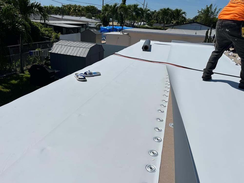 A worker installs white roofing material on a residential flat roof, showing a line of circular fasteners in the center.