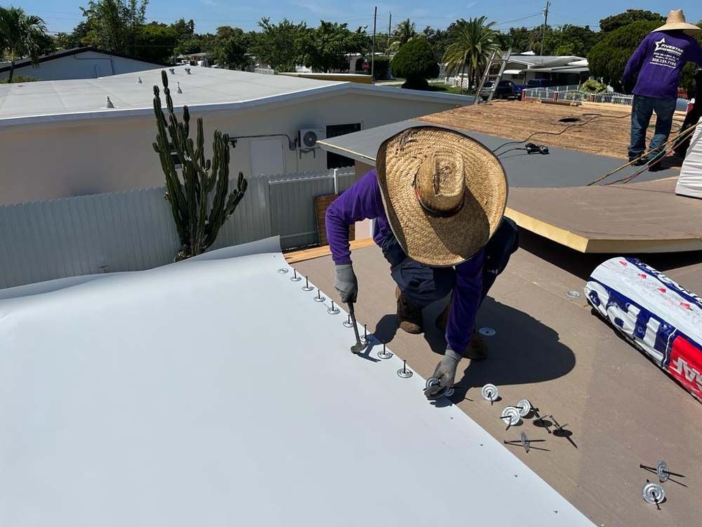 A worker in a straw hat installs fasteners on a white flat roof during a construction project.