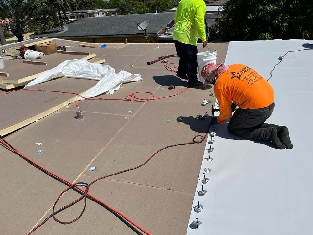 Two construction workers install a white roofing membrane on a flat roof, securing it with metal fasteners.