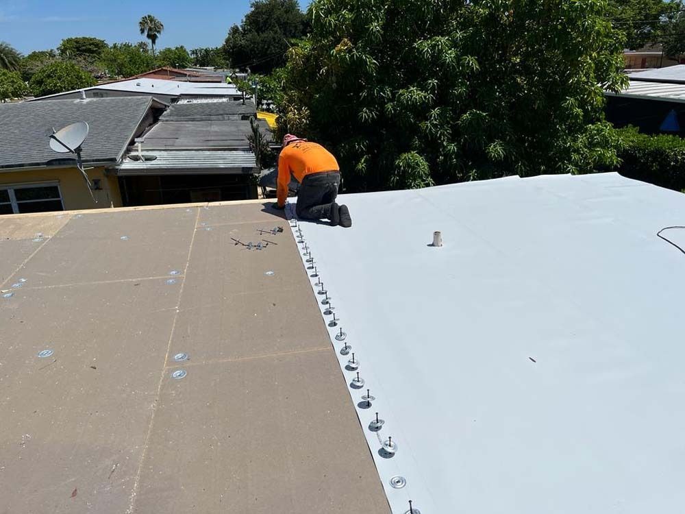 A worker in an orange shirt installs light gray roofing material on a flat roof, secured by a line of metal fasteners.
