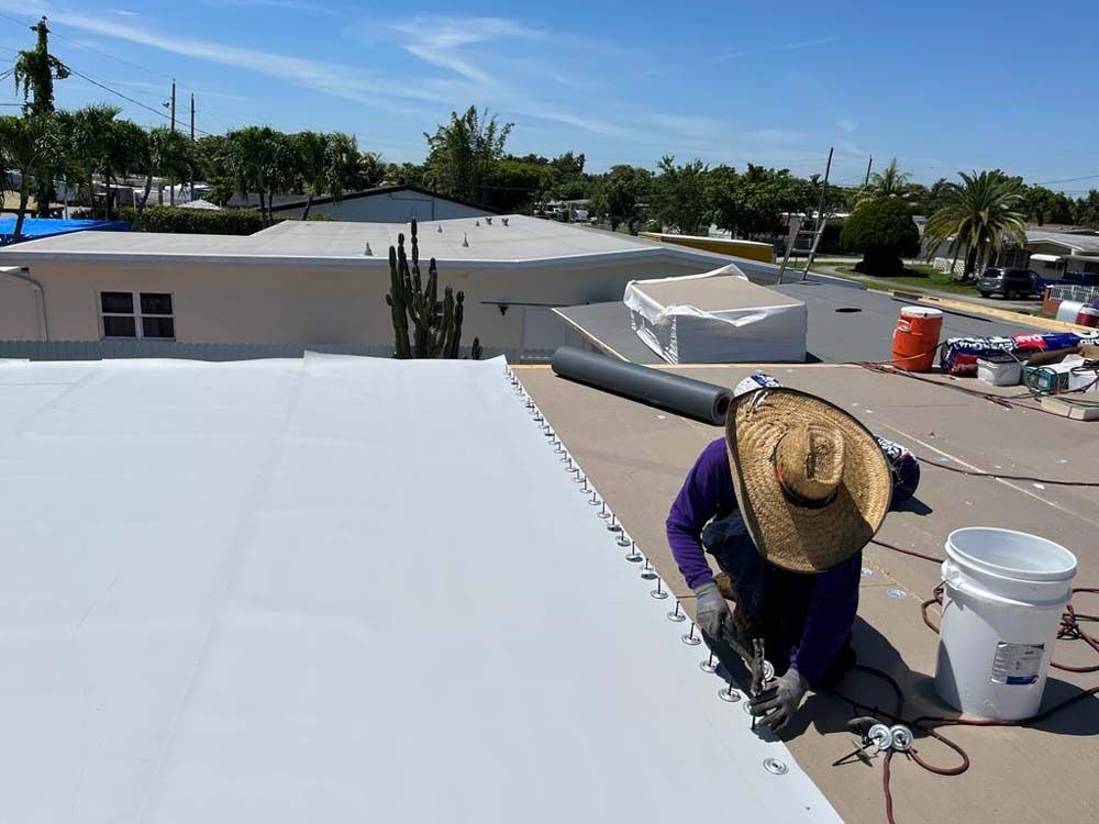 A person wearing a straw hat works on installing a white roofing membrane on a flat building roof under a blue sky.