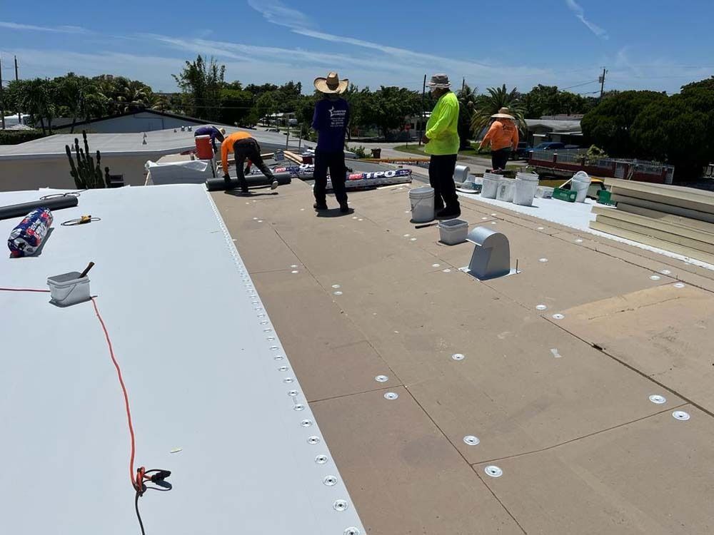 Workers install a white membrane roof on a sunny day, securing material with fasteners on the flat surface.