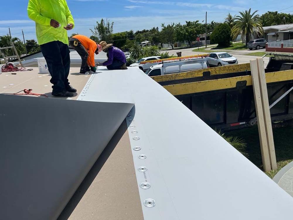 Three construction workers install a light-colored roofing membrane on an elevated flat roof on a sunny day.
