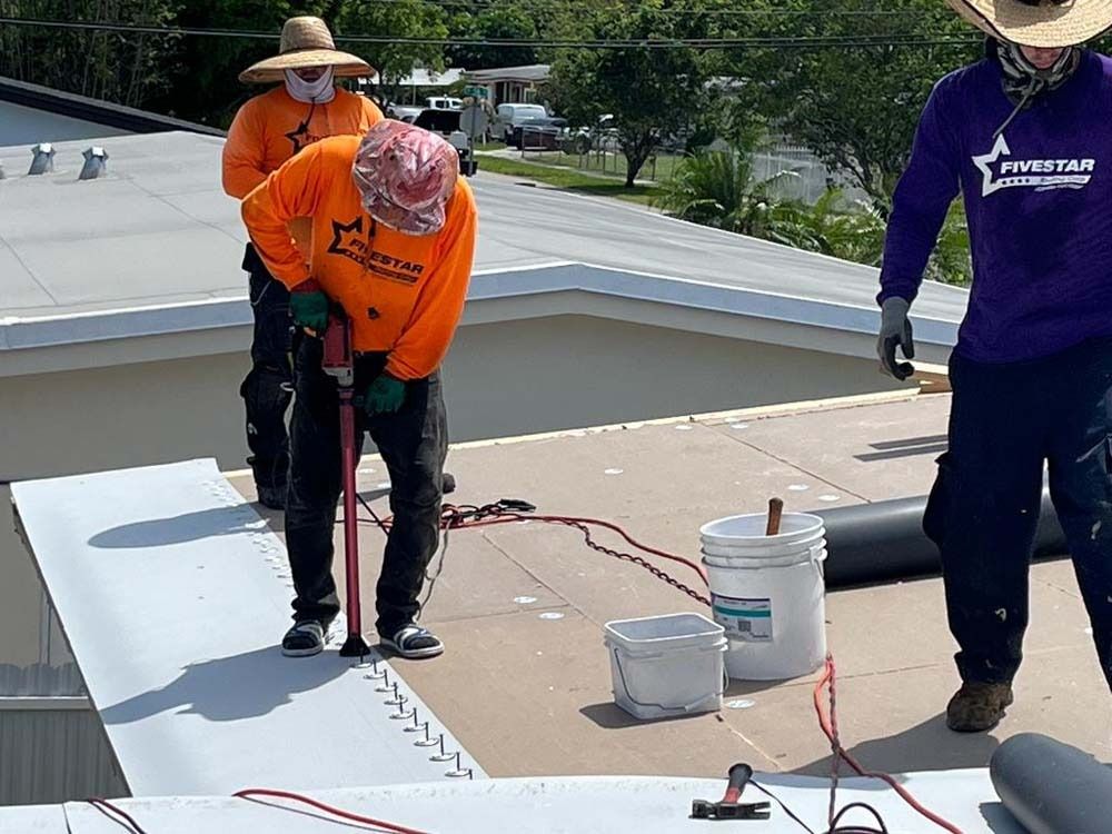 Three construction workers in long-sleeved shirts and hats install a light-colored roofing material on a flat roof.