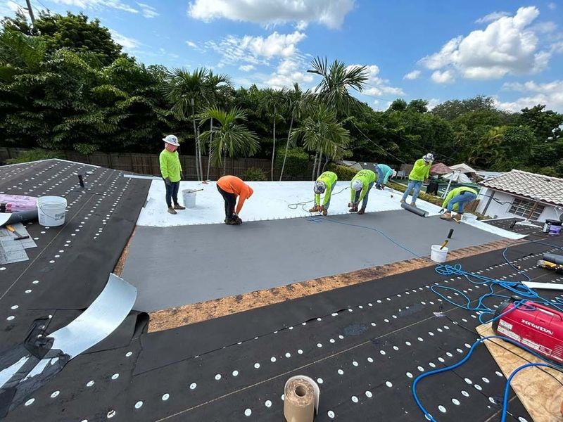 A crew in bright shirts and helmets works on a residential roof, installing white and grey roofing materials.