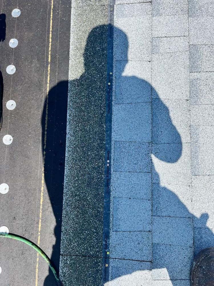 A person's shadow cast onto a dark curb and a light-colored paved walkway outdoors.