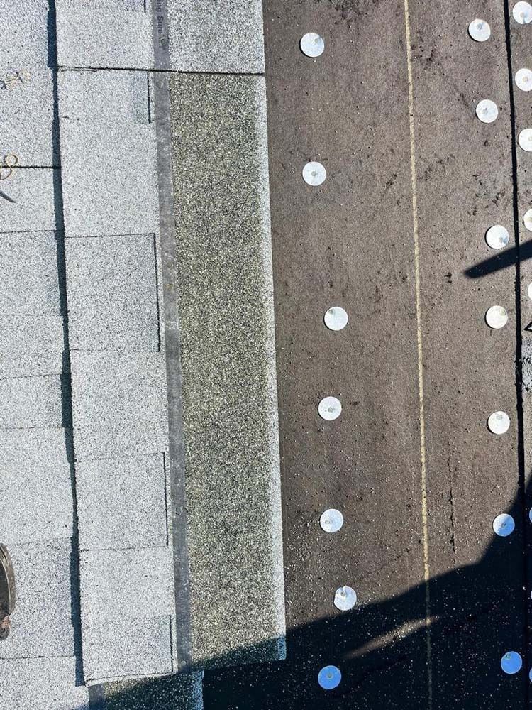 A close-up of roof shingles being installed over gray felt underlayment secured with evenly spaced metal cap nails.