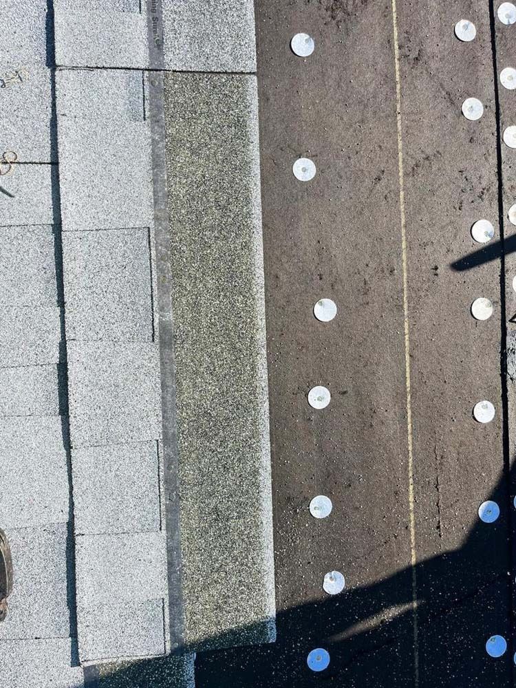 A close-up of a roof showing a vertical section of gray shingles next to dark underlayment secured with metal caps.