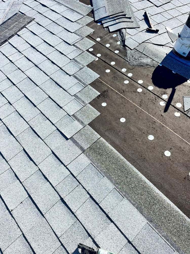 A partially shingled roof showing layers of asphalt shingles and rows of white circular fasteners on the underlayment.