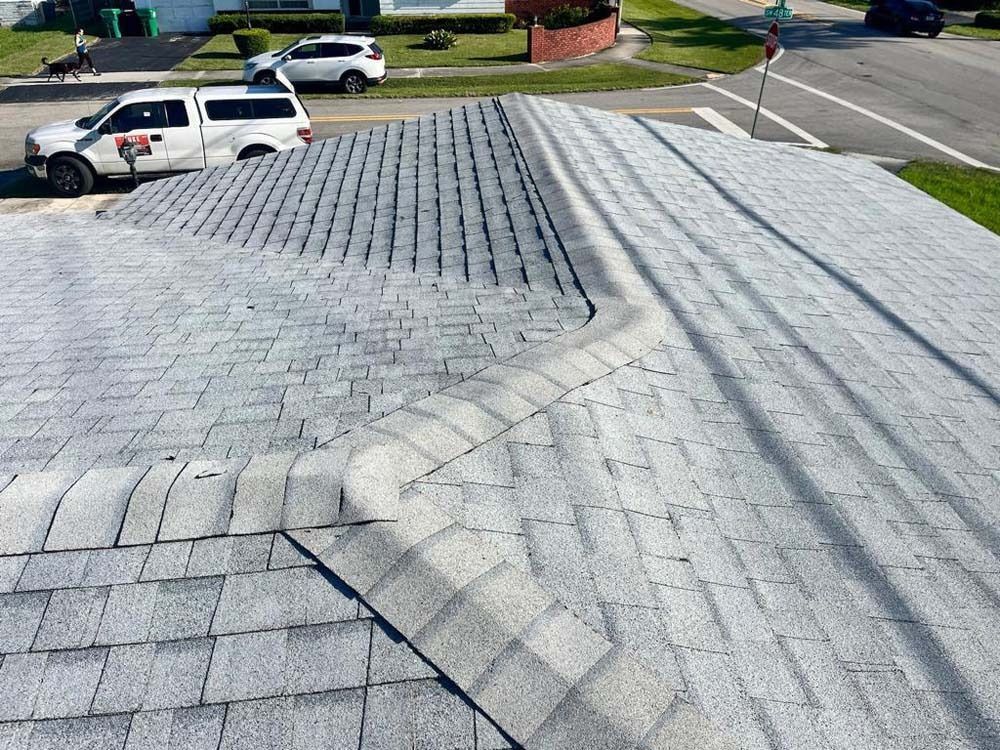 A high-angle view of a grey shingled roof with a complex hip and ridge line, with vehicles parked on a street below.