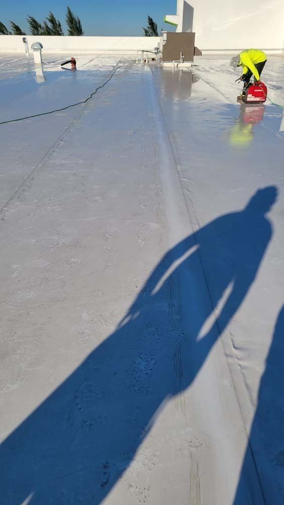 A person in a bright yellow safety vest uses a power tool on a light-colored flat roof on a sunny day.