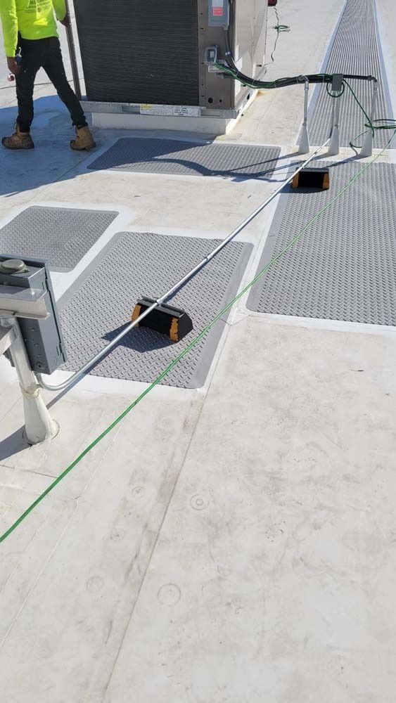 A worker in a high-visibility jacket walks near HVAC equipment on a flat rooftop with conduit supported by rubber blocks.