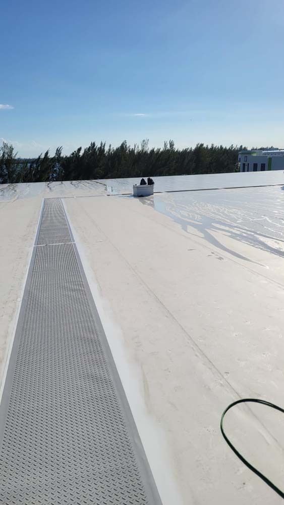 A view from a rooftop looking toward a worker on a flat, white roof near a line of trees under a clear, bright blue sky.