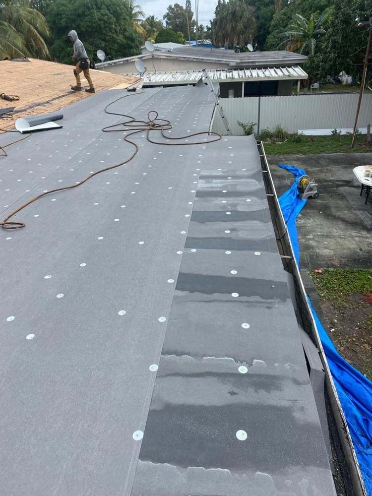A roofer works on a flat residential roof, installing gray roofing membrane secured with rows of white fasteners.
