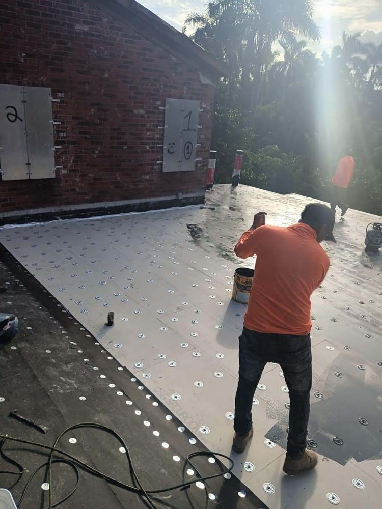 A worker in an orange shirt installs insulation boards onto a flat roof, securing them with numerous circular fasteners.