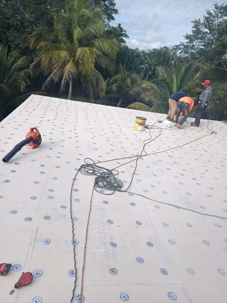 Three workers stand on a white roof surface marked with a grid pattern, surrounded by tropical trees.