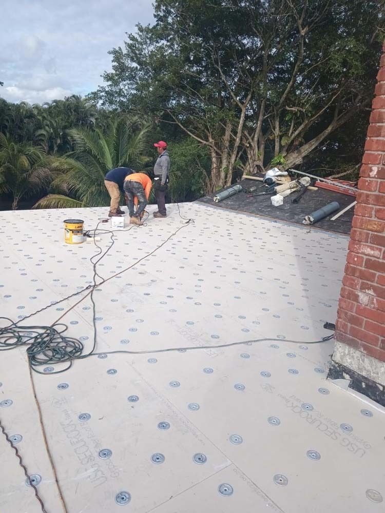 Three workers install insulation boards on a flat rooftop, next to a brick chimney and lush green trees.
