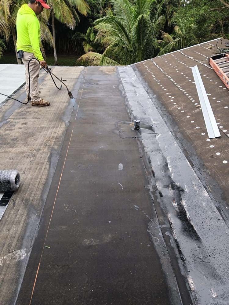 A worker in a high-visibility yellow shirt uses a torch to install roofing material on a sloped, shingled roof.
