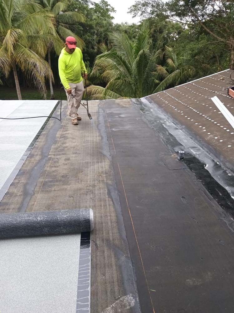 A person in a bright yellow shirt and cap applying roofing material to a sloped roof amidst palm trees.