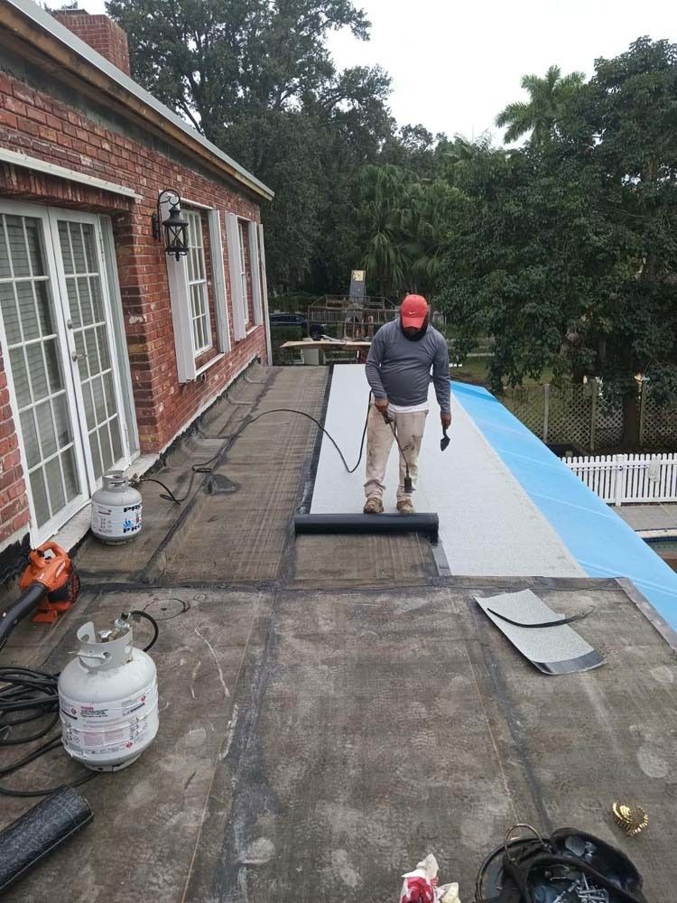 A construction worker in a hard hat uses a heat torch to install a roll of membrane roofing on a flat, brick house roof.