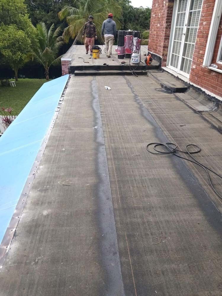 Two people work on a flat roof with asphalt rolls, near a brick wall and green trees.