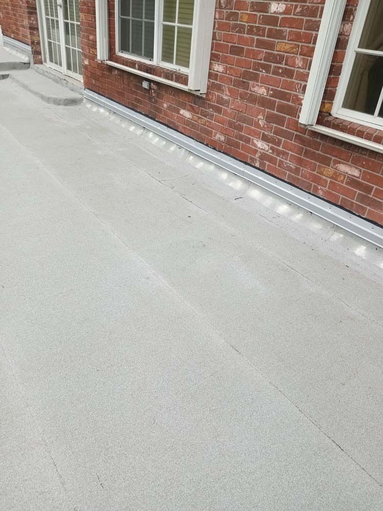 A grey concrete patio bordered by a brick wall with white-trimmed windows and a reflective metal edge trim.