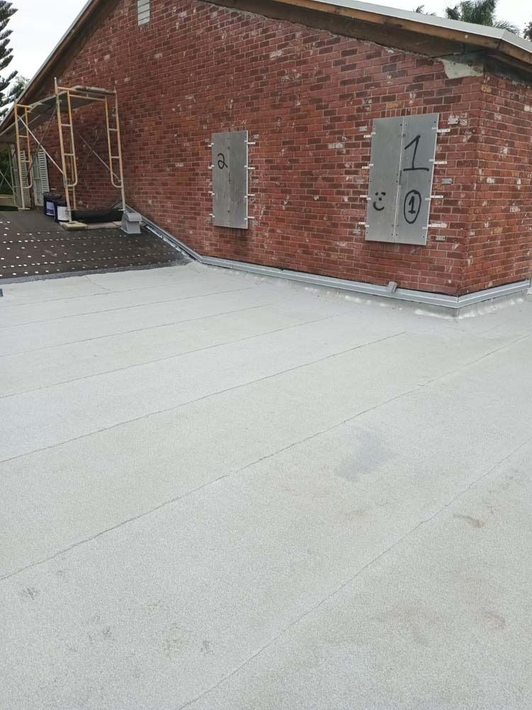 A red brick building wall featuring two boarded-up window openings above a newly installed light-gray flat roof.