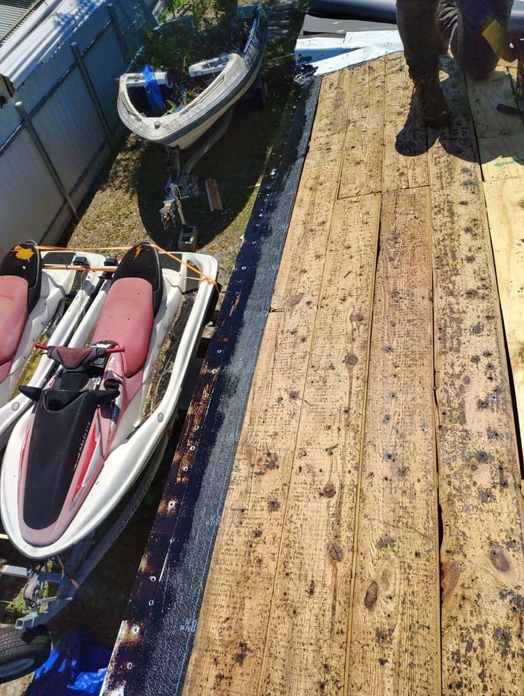 A person stands on a wooden deck next to two jet skis parked on a trailer beside a metal building.