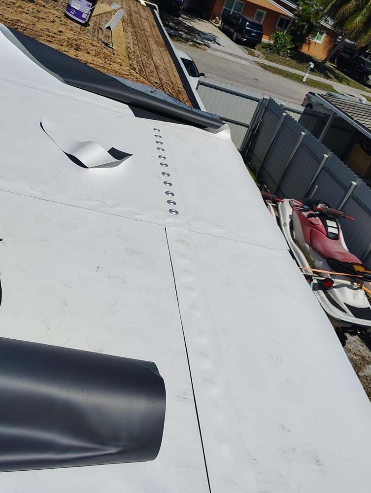 A high-angle view of a roof being repaired with white roofing material, showing seams and fasteners in a suburban setting.