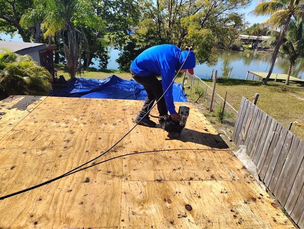A worker in a blue hoodie uses a nail gun to fasten plywood sheets onto a residential roof by a lake.