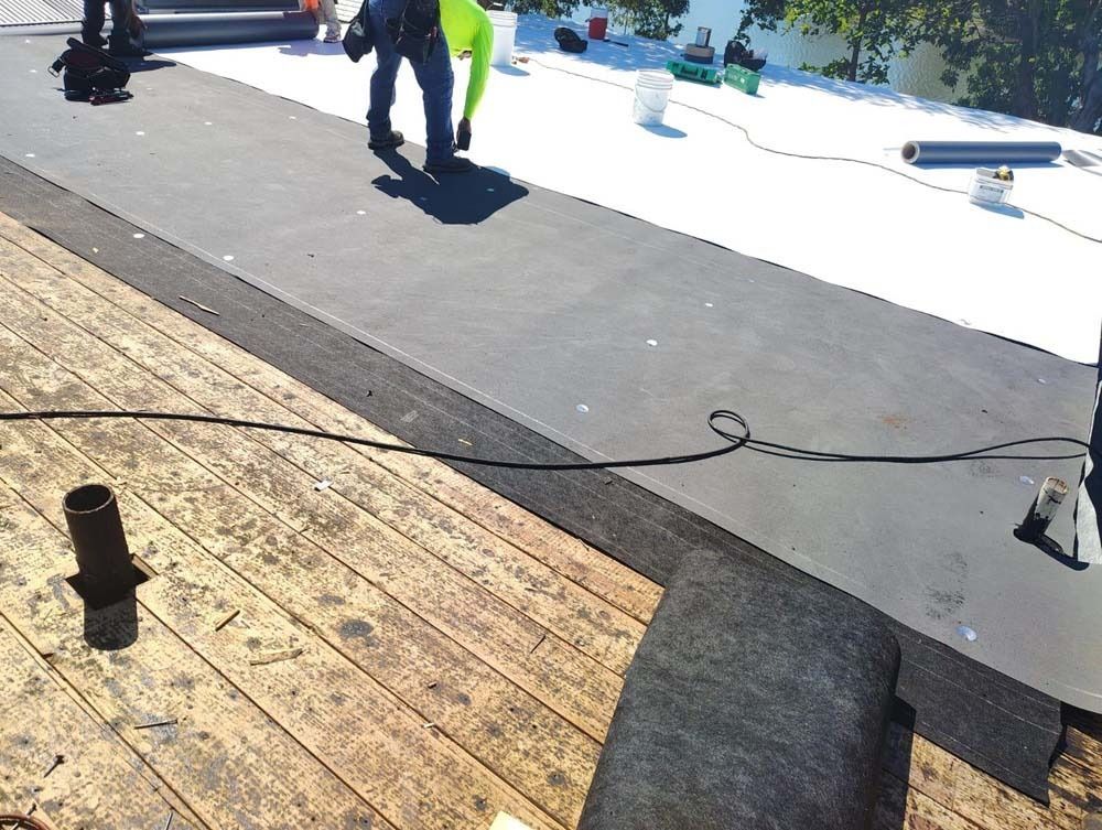 A roofer installs black roofing membrane on a wooden substrate next to a white surface on a sunny day.