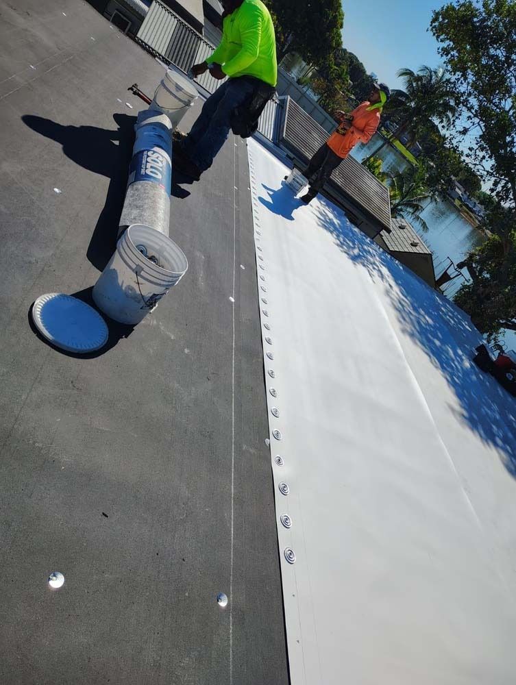 Two workers apply a white coating to a flat, dark roof surface, with a bucket and supplies visible nearby.