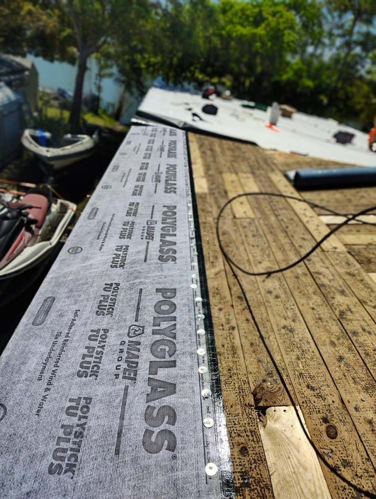 A roll of Polyglass underlayment being installed on a wooden roof deck near a lake, with tools visible in the background.