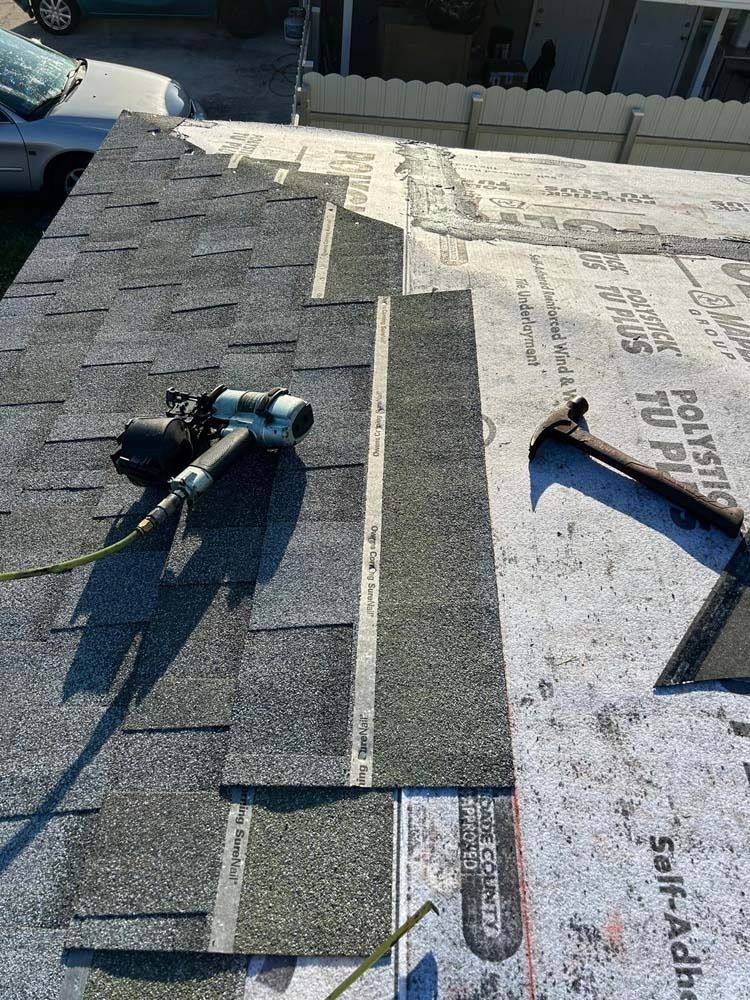 A partially shingled roof with a nail gun, hammer, and underlayment rolls visible in a residential setting.