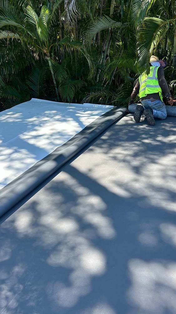 A worker in a high-visibility vest installs rolls of white roofing membrane on a flat roof surrounded by palm trees.