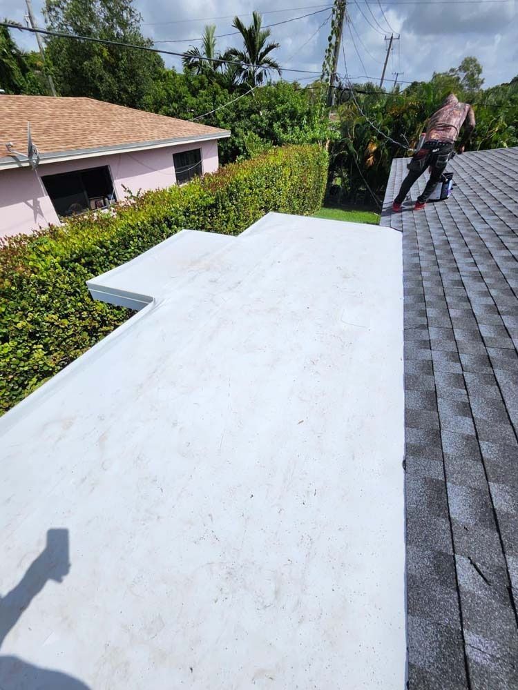 A worker stands on a gray shingled roof next to a freshly coated, light-gray flat roof section on a sunny day.