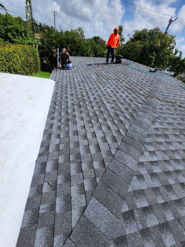 Two workers in high-visibility orange clothing install gray asphalt shingles on a sloped residential roof.
