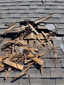 A large hole in a shingled roof with splintered wooden debris scattered around the opening.
