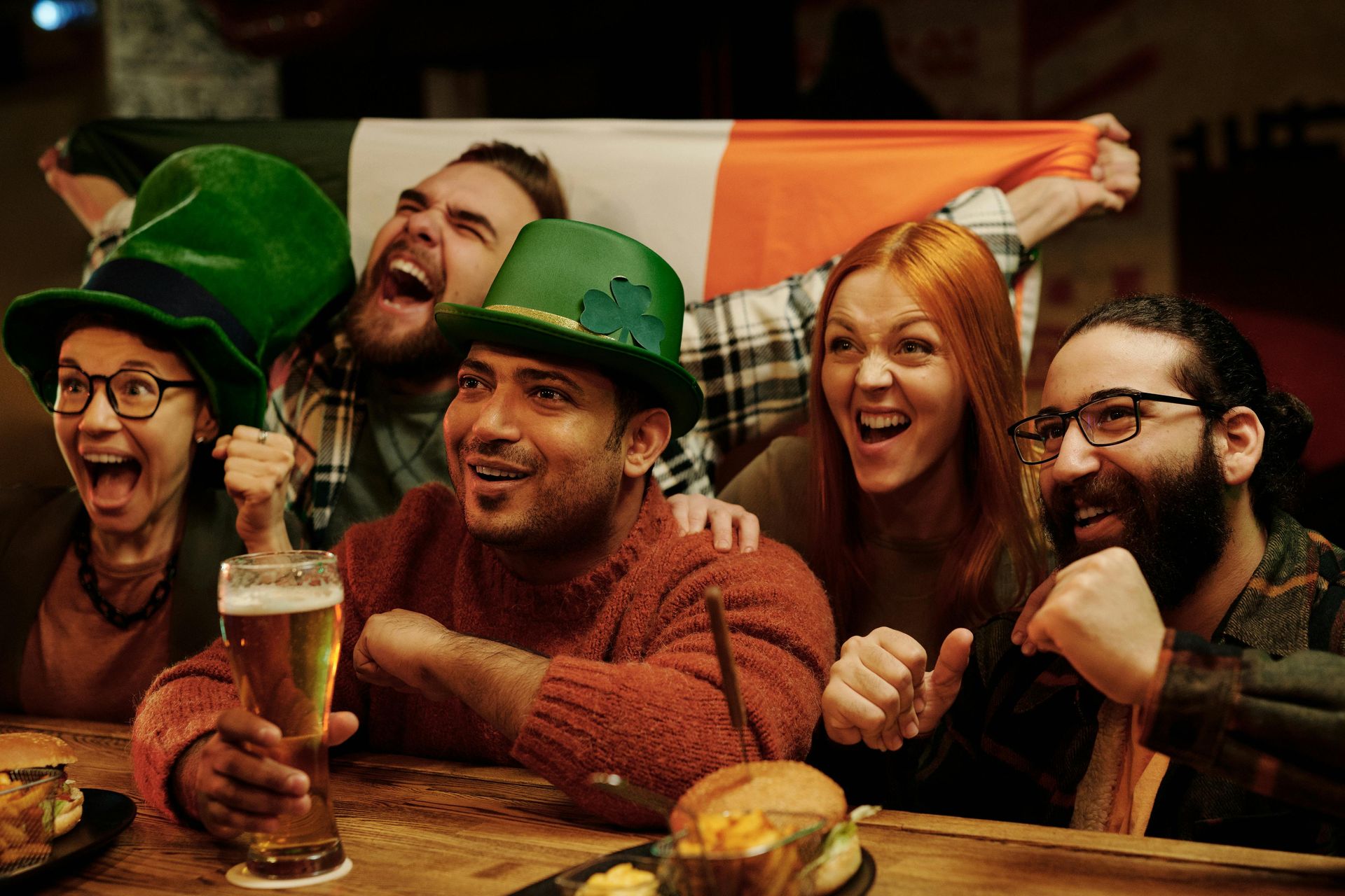 Group of friends celebrating in a pub, some wearing green hats and holding an Irish flag, cheering with beer.