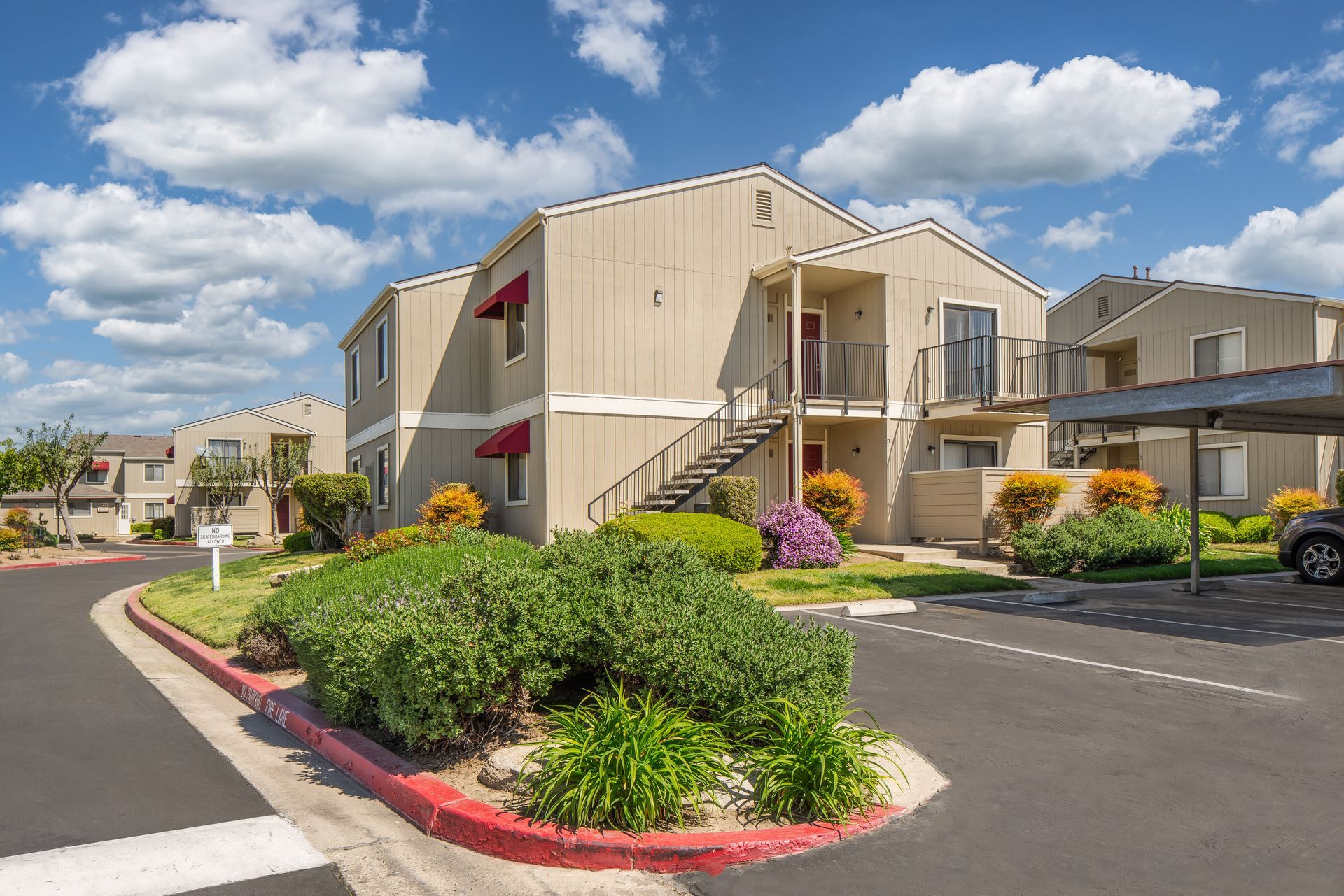 Apartment complex exterior with beige buildings, stairs, red awnings, lush landscaping, and a car park, under a blue sky.